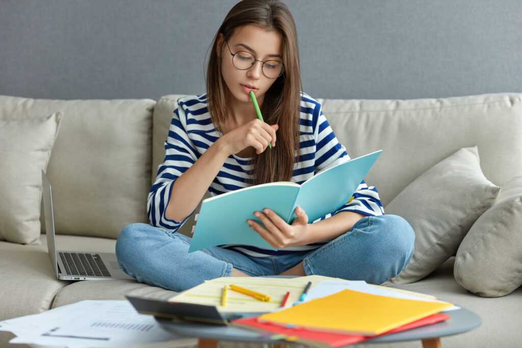 studying online concept serious young woman being busy with remote freelance project sits at comfortable sofa writes notes holds textbook use laptop computer at home with wireless internet